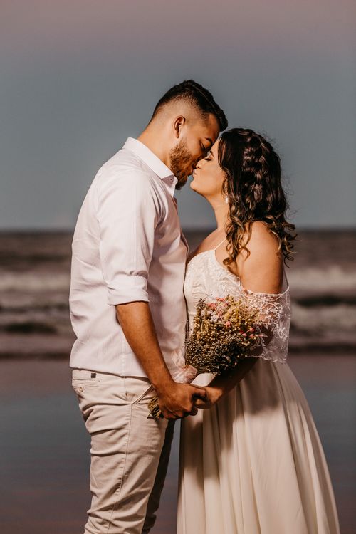 Free Side view young ethnic couple wearing elegant white wedding gowns holding hands and kissing tenderly while standing against blurred waving sea Stock Photo