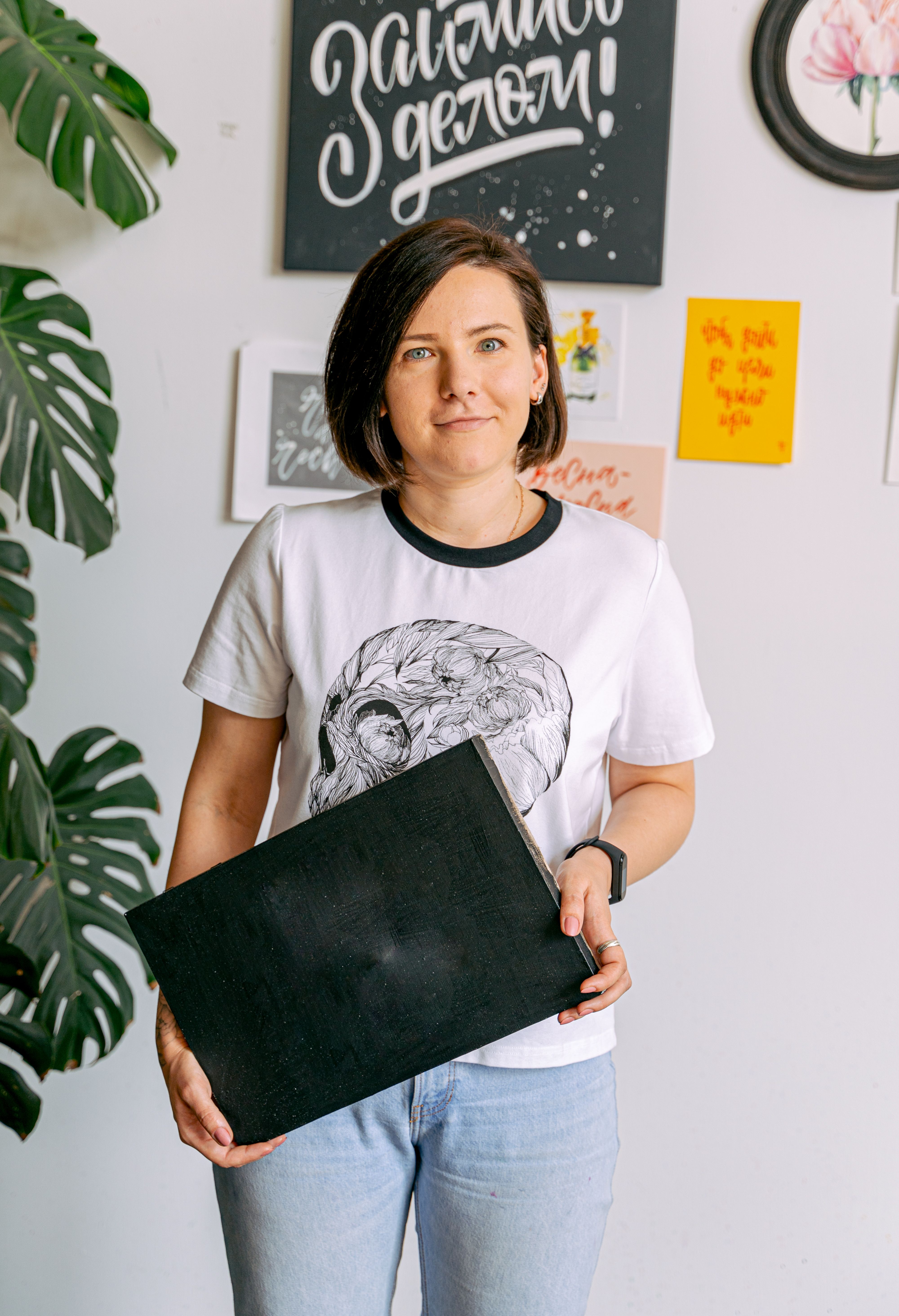 Caucasian woman holding a blank black canvas standing indoors, with art on the wall.
