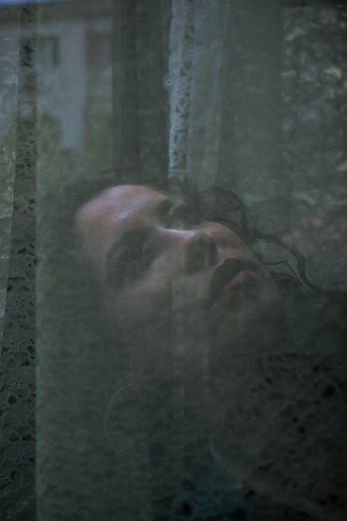 Double exposure of thoughtful female with curly hair standing near window with curtains and looking up