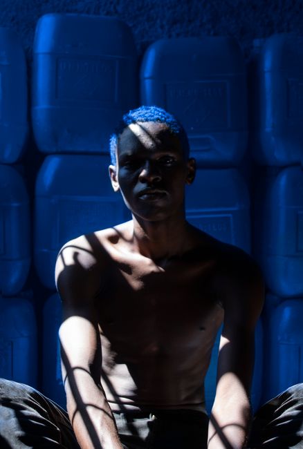 Dramatic portrait of a shirtless man sitting in shadows against blue jerry cans.