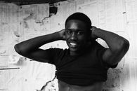 Black and white of cheerful provocative male standing near paper wall and taking off t shirt while looking at camera