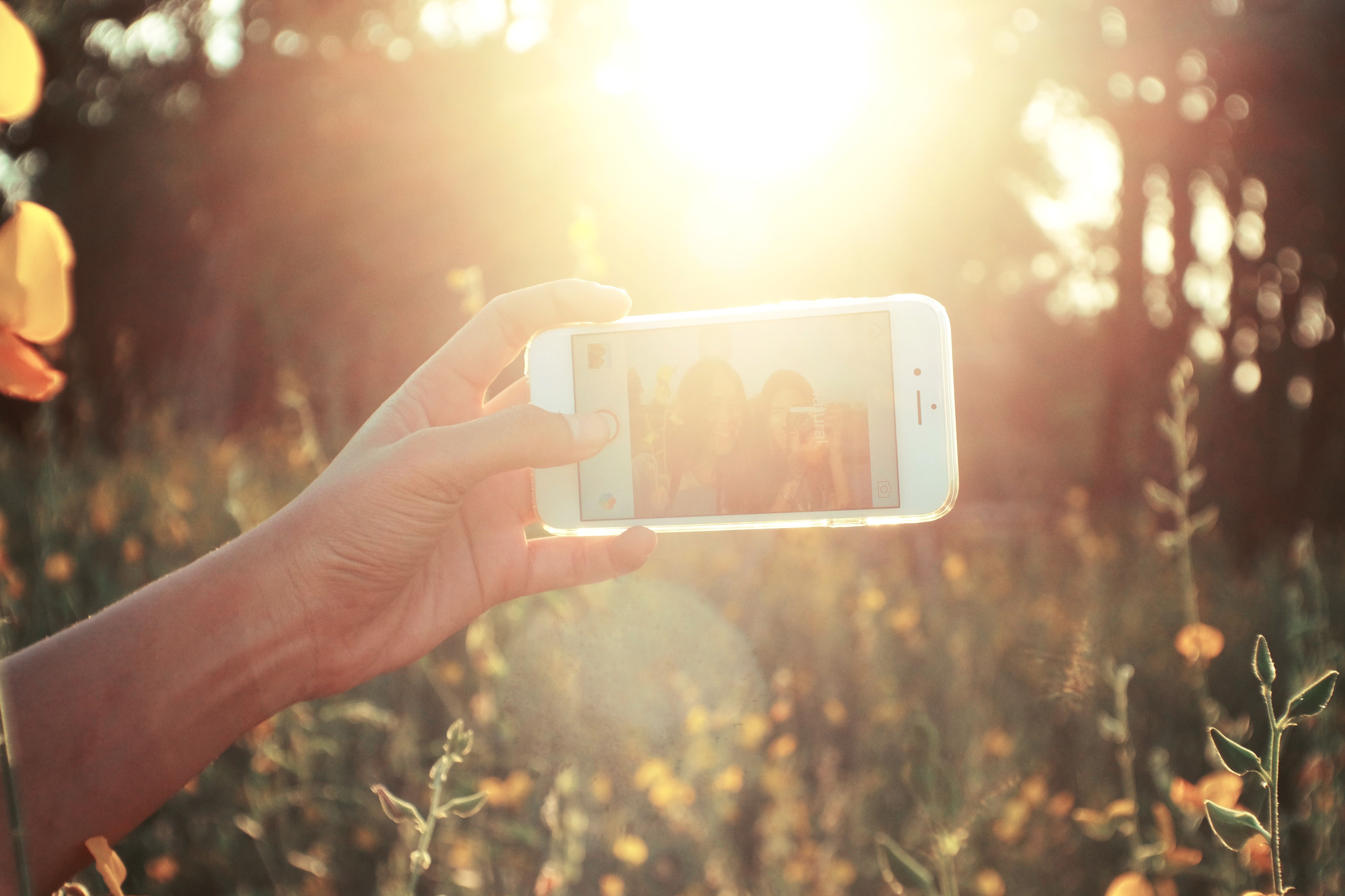 A group of friends laughing together outdoors on a sunny day while one holds a cell phone.