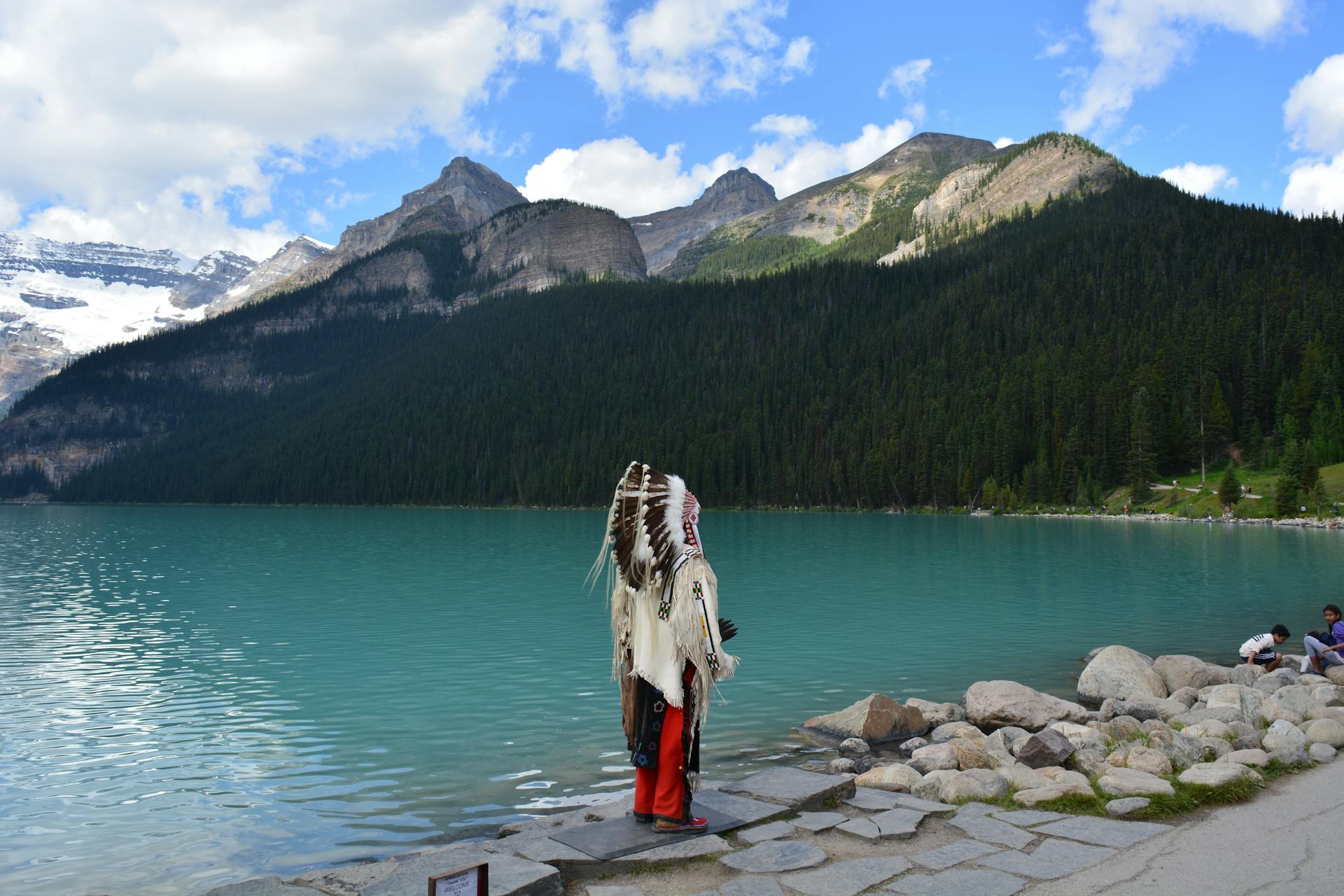 Indigenous man in traditional headwear stands by Lake Louise with stunning mountain backdrop.