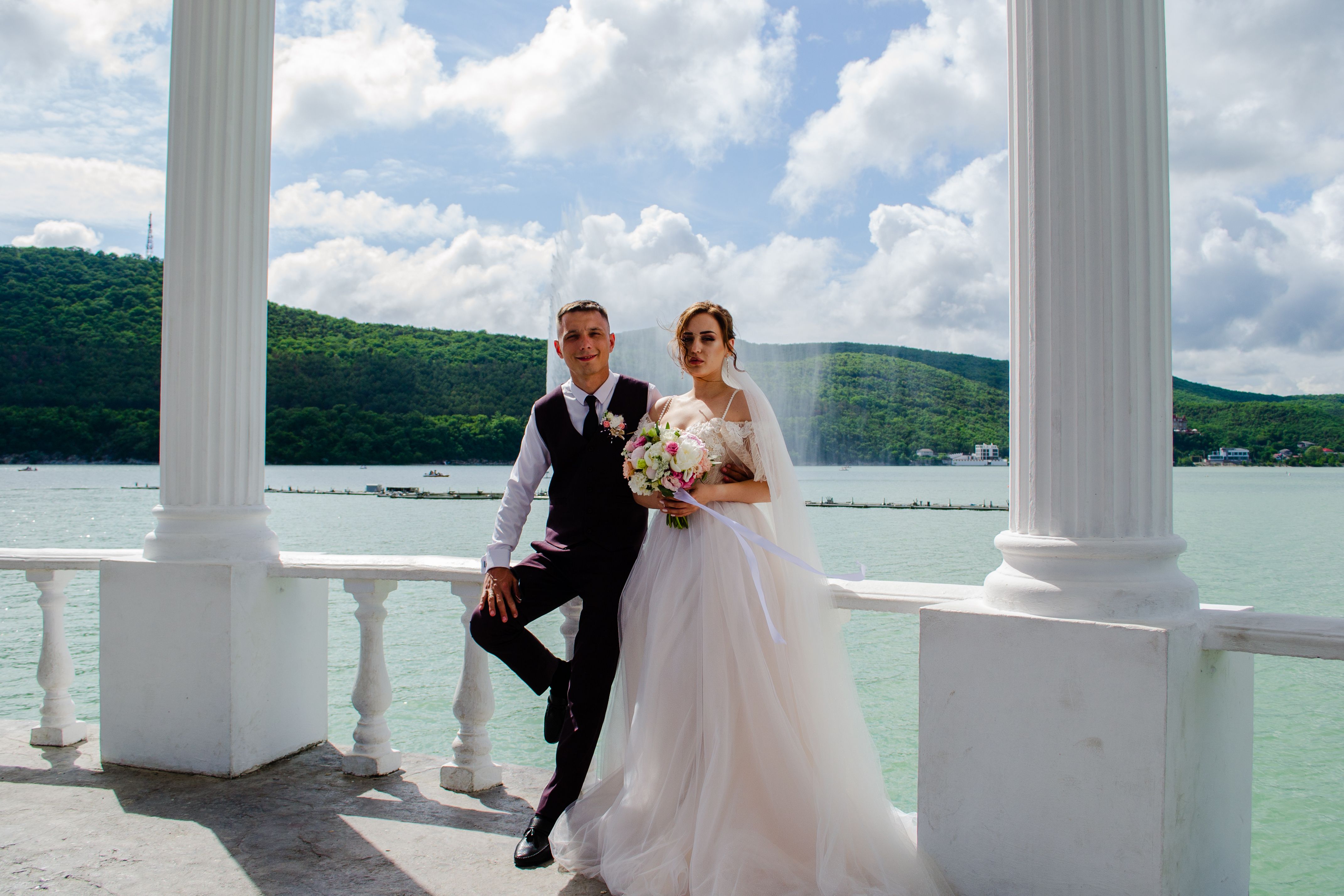 A newlywed couple poses elegantly by a picturesque lakeside, under clear skies.