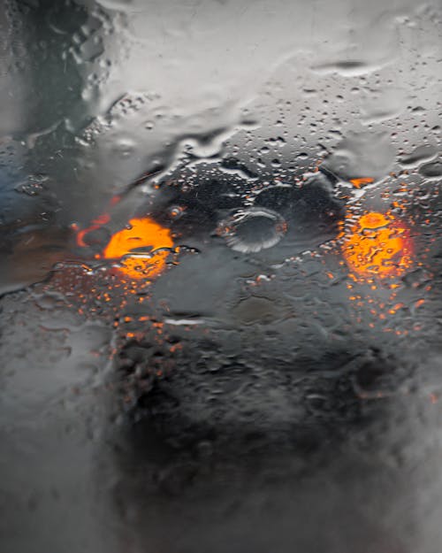 Wet glass with rain drops and spots located in front of blurred modern car with glowing orange headlights