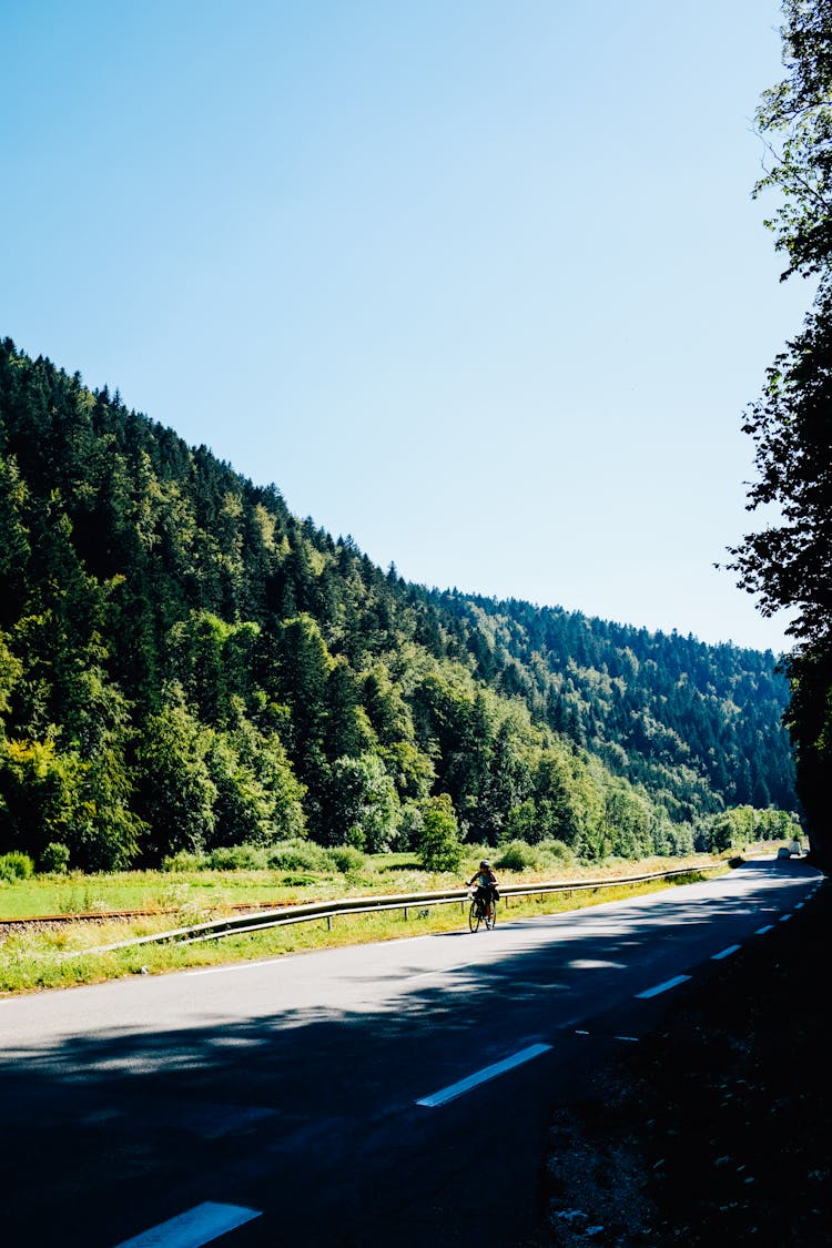 Faceless Cyclist Riding On Road Near Green Meadow And Mountain