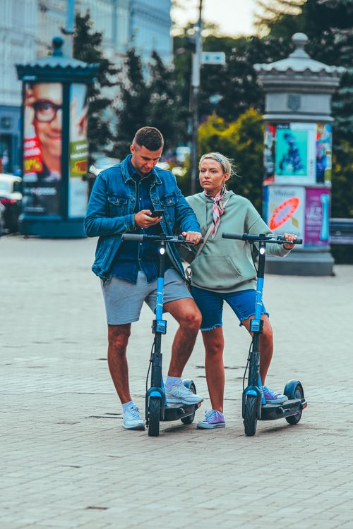 Free Full body of adult man scrolling smartphone while standing neat woman with scooter in city Stock Photo