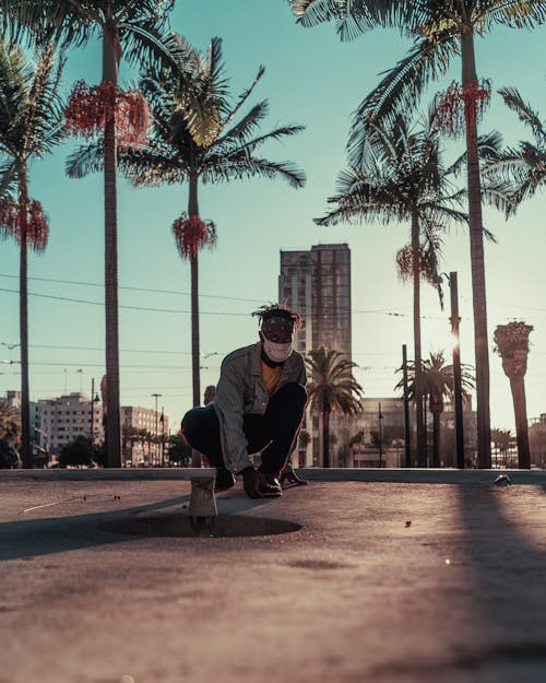 A Man in Denim Jacket Wearing Face Mask Sitting on the Ground Near Palm Trees while Looking at the Camera