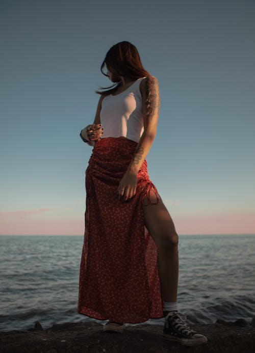From below full body of anonymous female in long skirt and top admiring rippling sea under bright sundown sky
