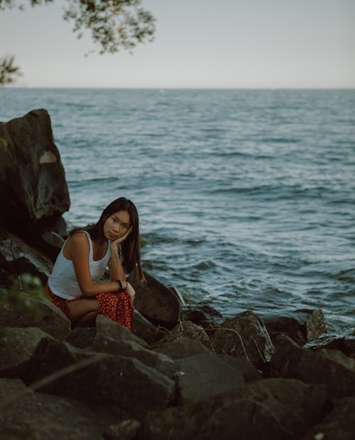 Pensive young Asian female leaning on hand while sitting on stone near rippling sea and looking at camera