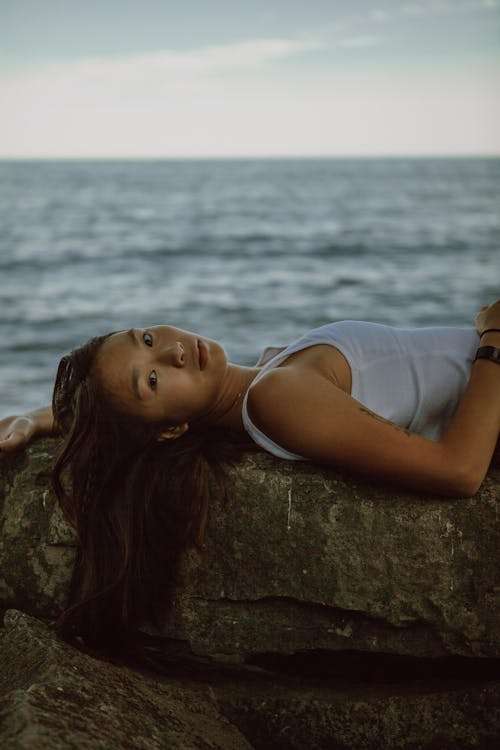Side view of relaxed Asian female enjoying rest and lying on stony coast near rippling sea