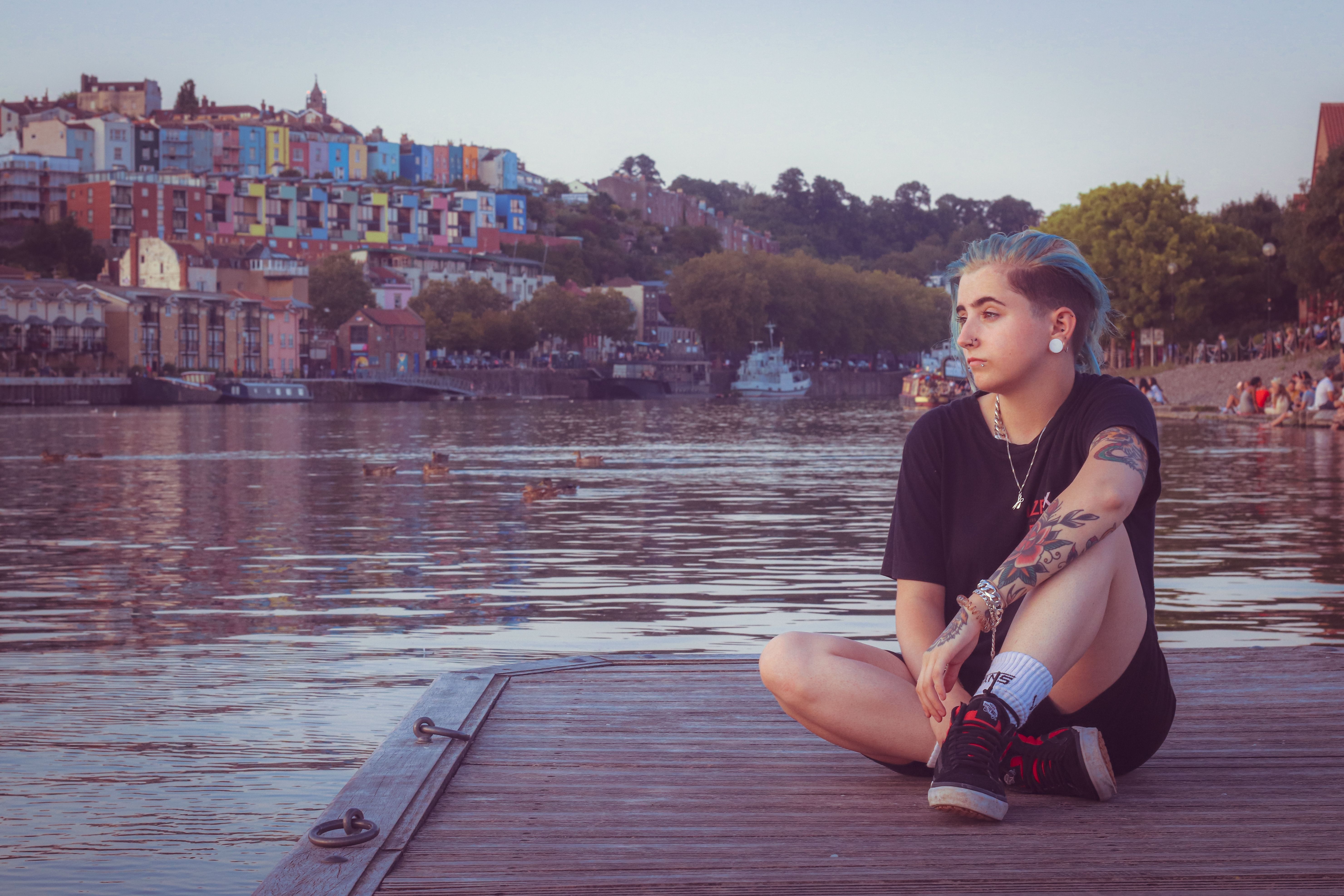 Full body of focused female with short hairstyle and tattooes sitting on wooden pier near river water and city buildings