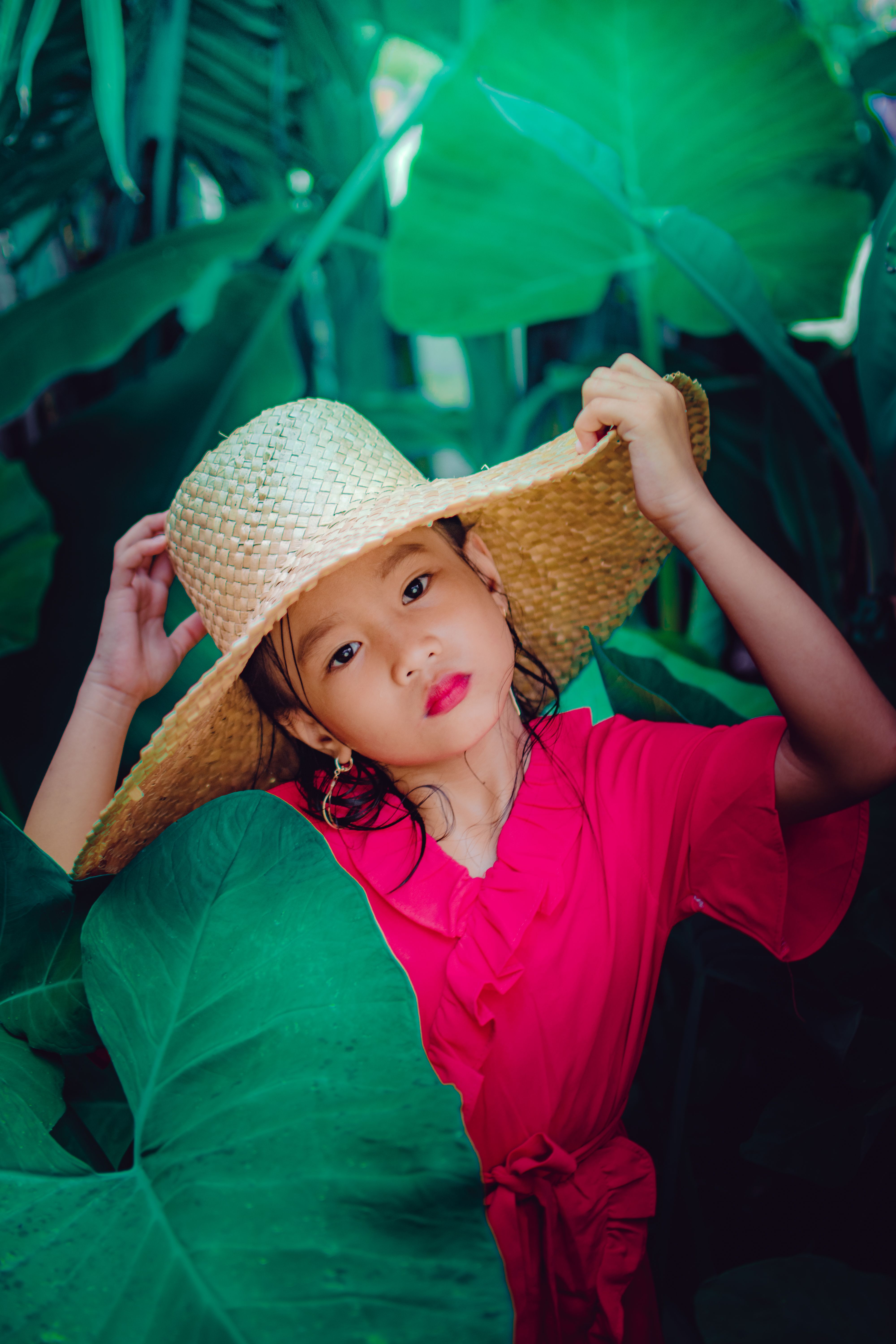 一位头戴草帽、身穿粉色连衣裙的年轻女孩，在郁郁葱葱的绿植间摆好姿势，捕捉着户外宁静的一刻