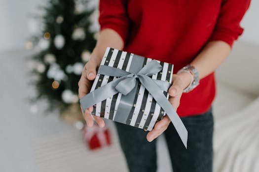 Close-up of a person holding a wrapped gift box with a festive ribbon, in front of a Christmas tree.