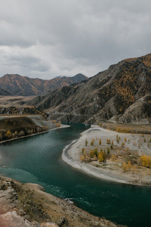 Picturesque view of stream flowing between mountains with plants under cloudy sky in winter