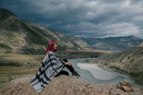 Side view of stylish female in warm scarf relaxing on rocky hill near mountains and river with ice