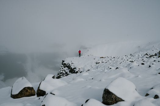 一位孤独的冒险家身着红色夹克，站在冰岛白雪皑皑的悬崖上，四周雾气弥漫