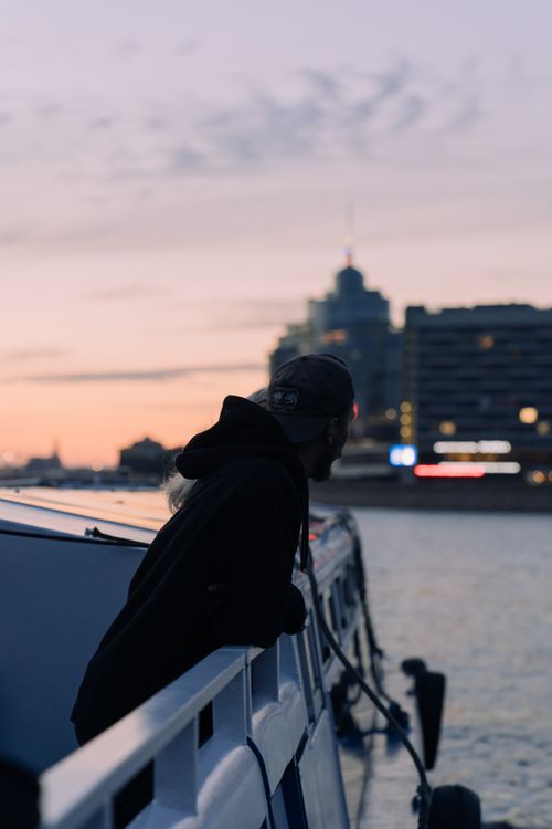 Free Man in a hoodie gazes over a city waterfront at sunset, blending urban tranquility. Stock Photo