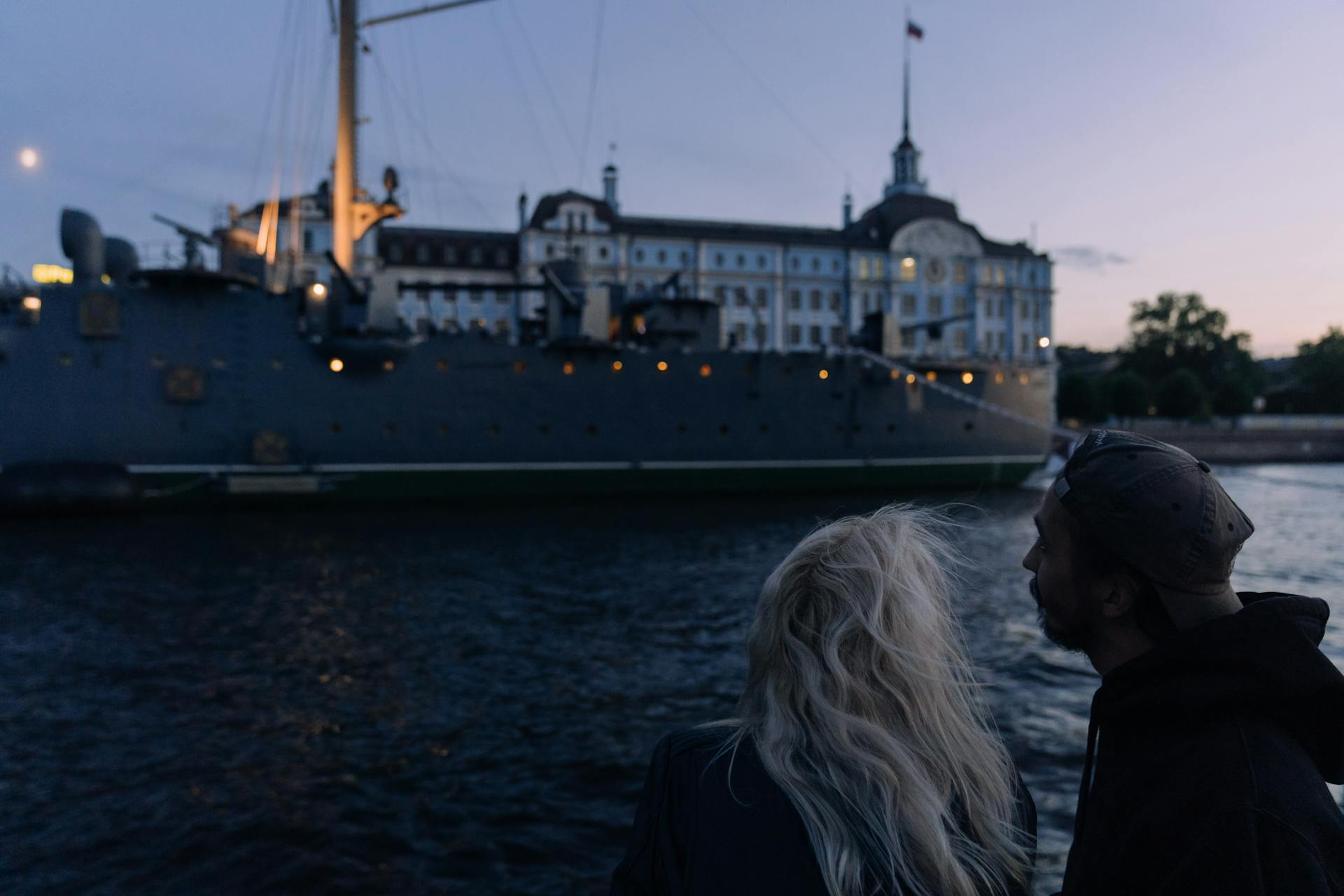 A couple gazes at a historic warship docked by a grand building at dusk, creating a romantic evening scene.