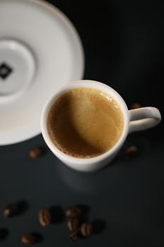 A close-up view of a freshly brewed espresso shot in a white cup with coffee beans around it.