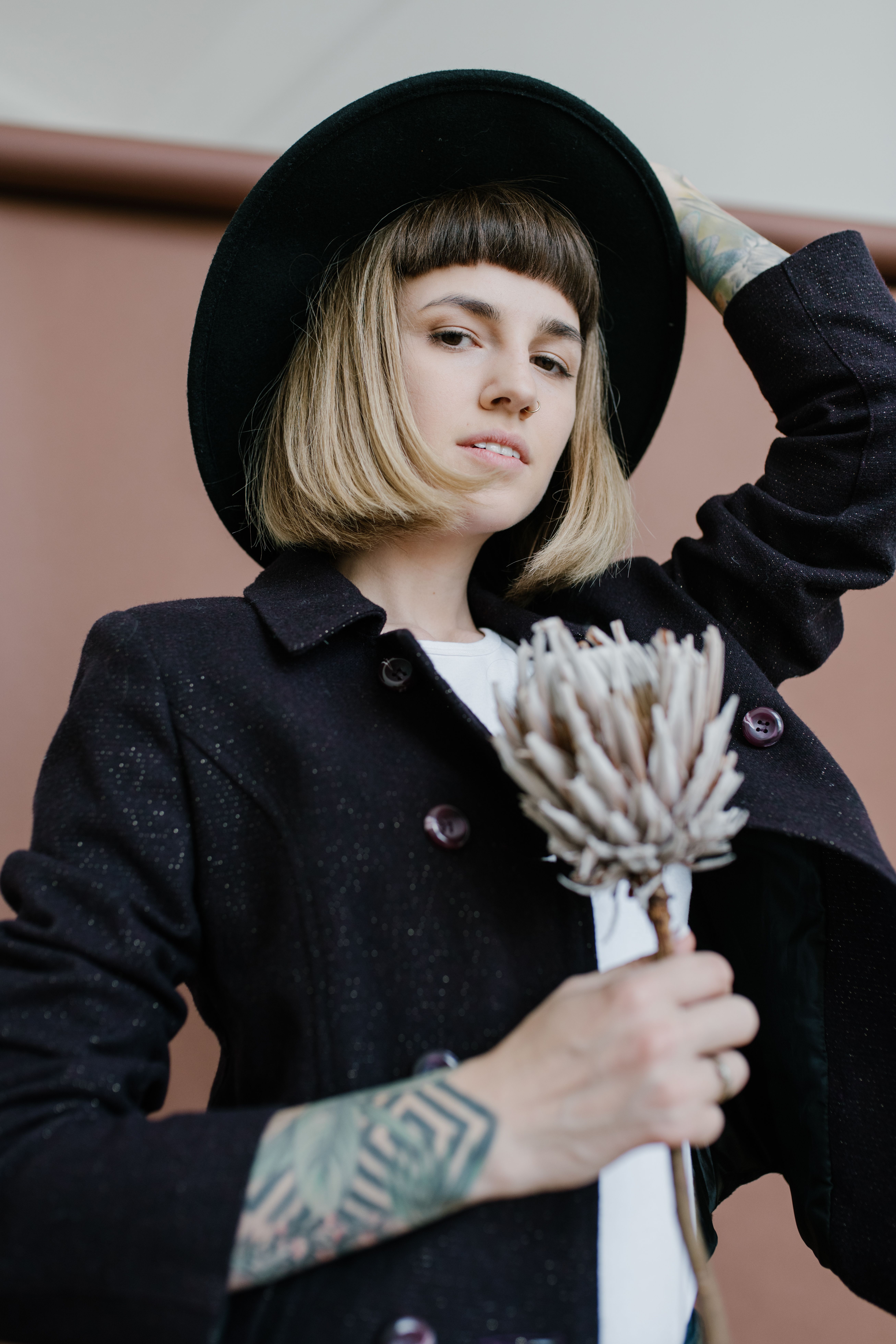 Low angle of tattooed female in hat holding bunch of flowers and looking at camera