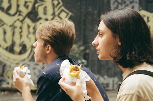 Two young adults savoring sandwiches outdoors, depicting a casual urban scene.