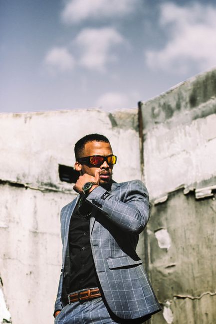 A stylish man in sunglasses and formal attire poses confidently against an outdoor backdrop.