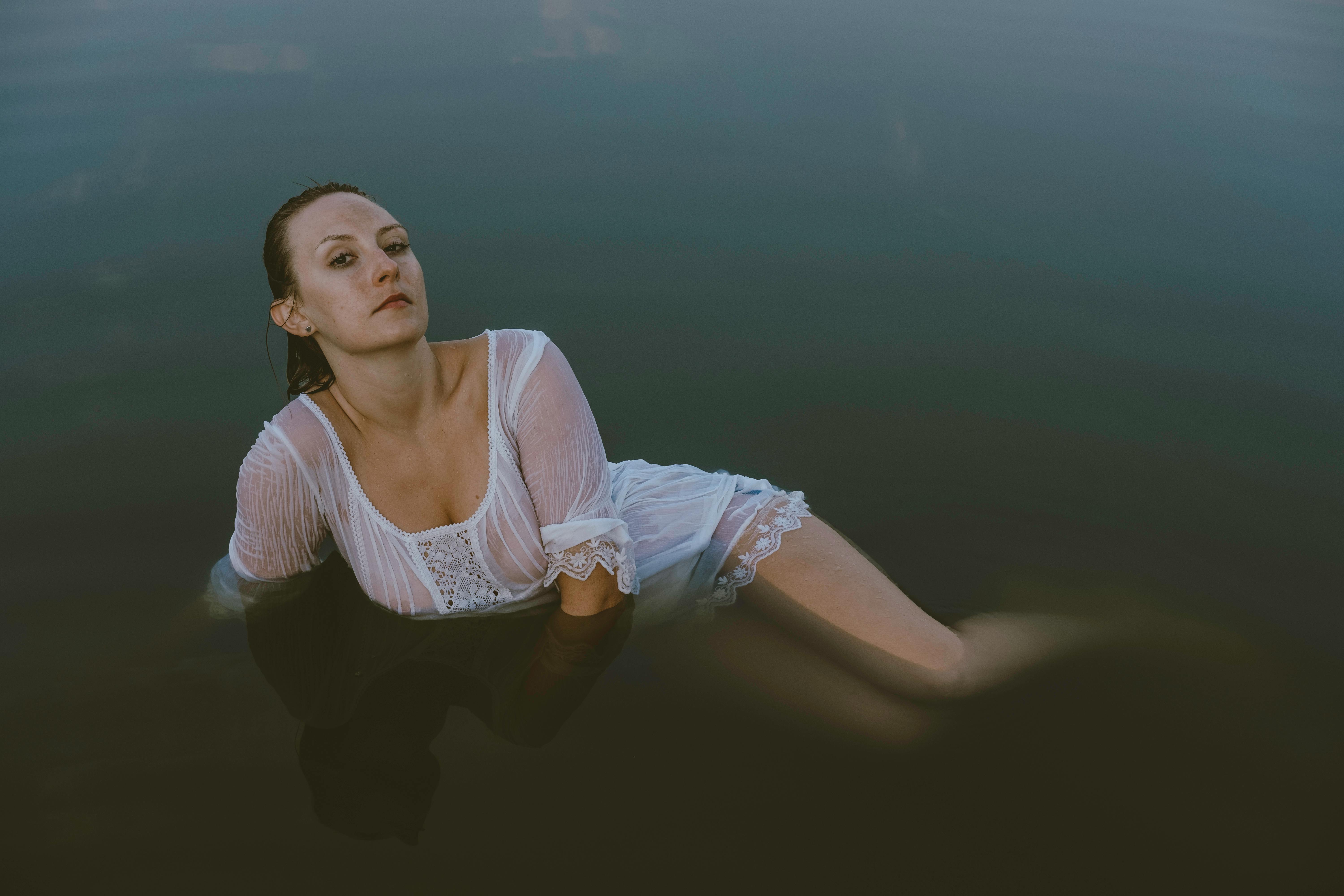 Full body of young beautiful female with wet hair in short white dress looking at camera while chilling in pond near coast