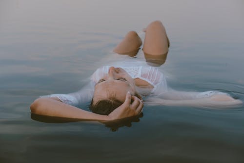 Full body of young slender female in short white dress touching wet hair while resting in calm lake in daylight