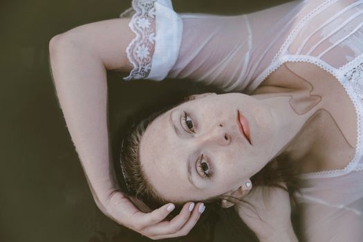 From above of serious female in transparent blouse bonding hand around head while lying in water and looking at camera