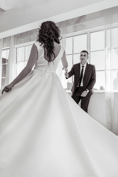 Black and white of unrecognizable fiancee in long white bridal gown spinning and holding hand of groom in elegant tuxedo in bright room