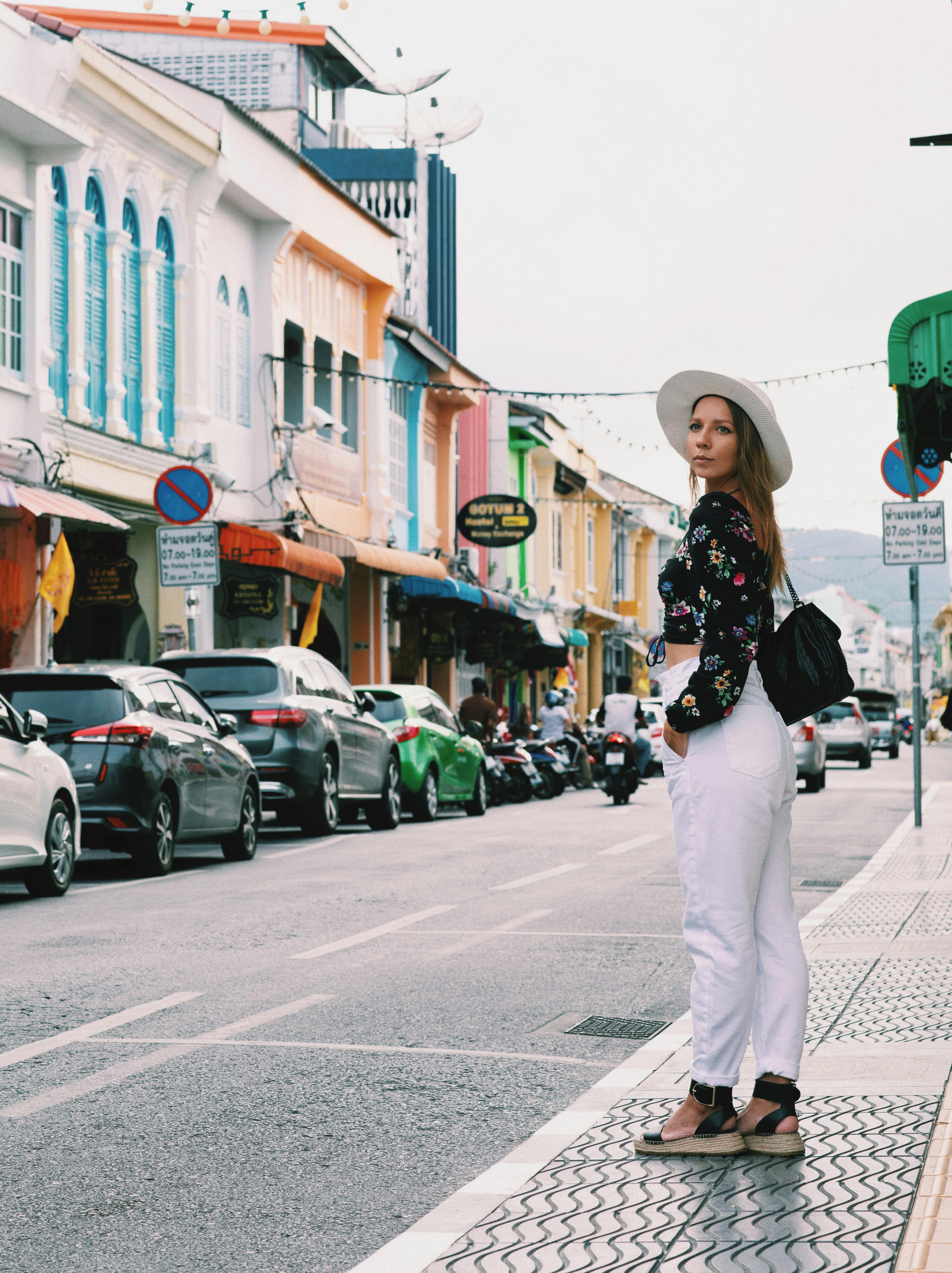 a woman in black floral crop top and white pants standing on the side of the road while looking afar