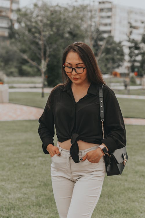 Young pondering Asian lady in stylish apparel and eyeglasses standing with hands in pockets on lawn while looking down in town