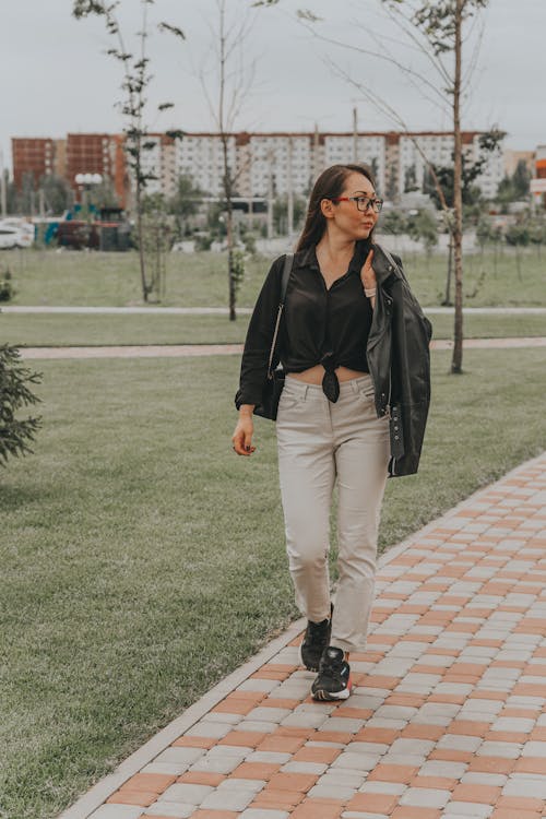 Full body of young black haired female in trendy clothes and glasses walking on paved walkway in park and looking away
