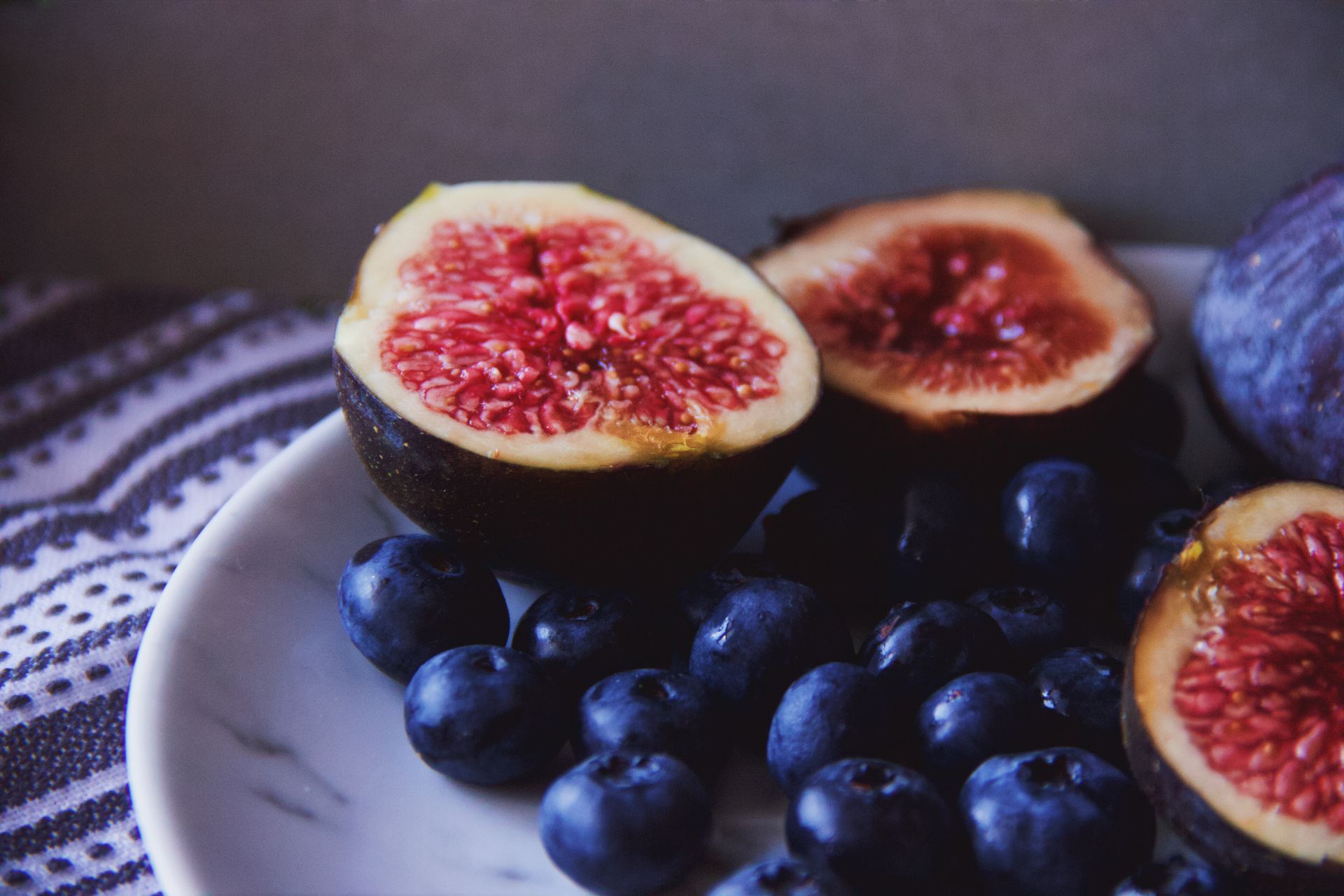 Halves of ripe figs placed on plate near blueberries on table with cloth in kitchen in daylight inside
