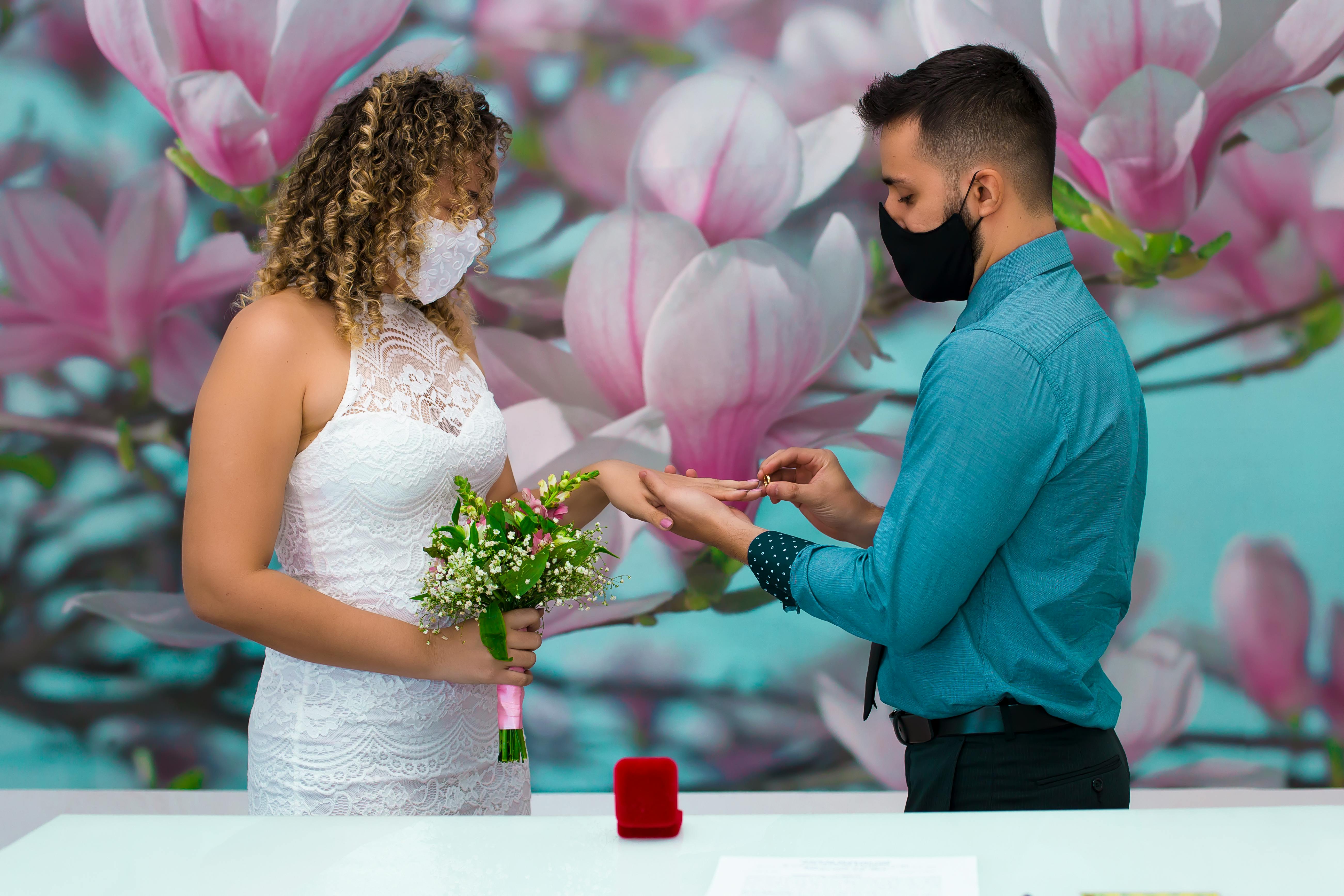 A couple exchange rings wearing masks during an intimate wedding with floral backdrop.