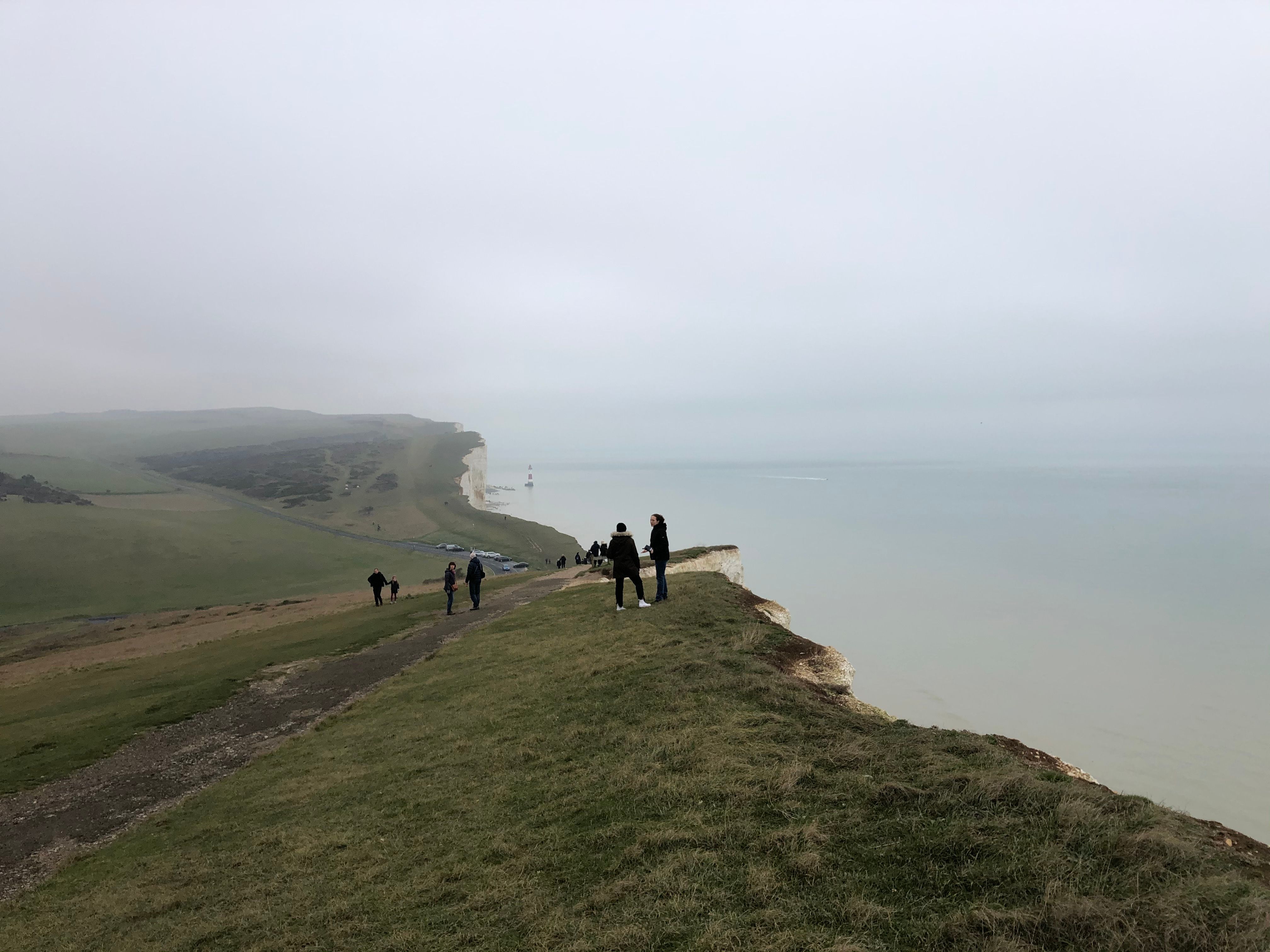 Tourists stand on the famous Dover cliffs overlooking the calm sea under a cloudy sky.