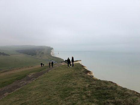 Tourists stand on the famous Dover cliffs overlooking the calm sea under a cloudy sky.