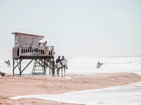 People enjoying a summer day by the ocean with surfers and a lifeguard tower.
