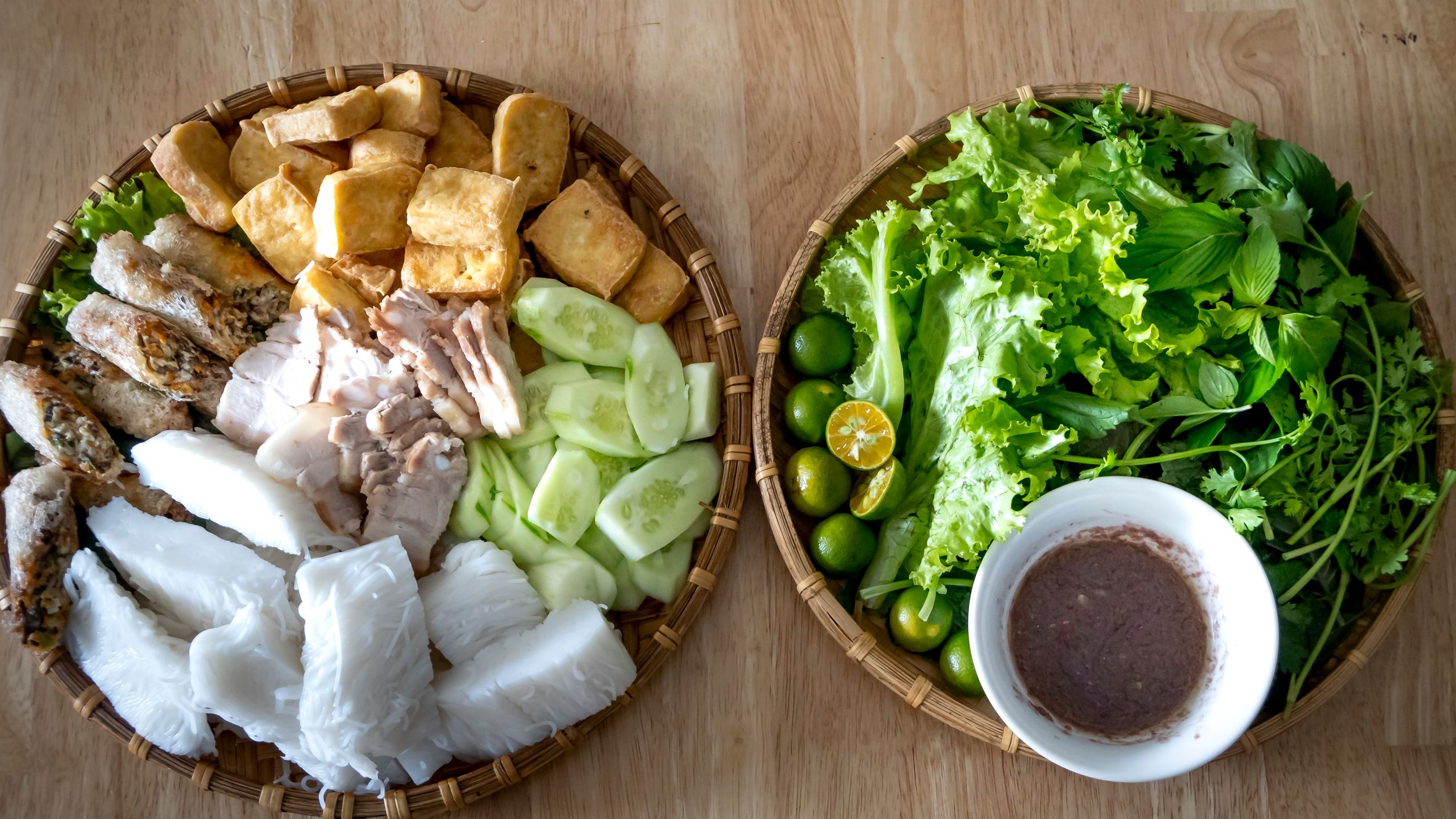 Top view appetizing traditional Vietnamese dish with fried tofu cut cucumbers and boiled noodles served in bowl on table near bowl with mixed green leaves and ripe lime