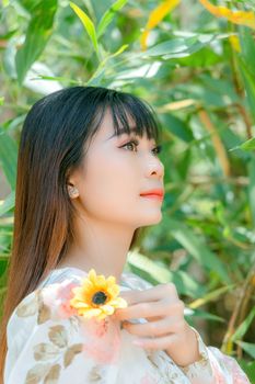 A serene portrait of a young woman holding a yellow flower, surrounded by lush greenery.
