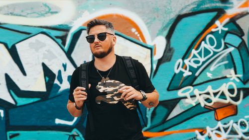 Free Adult male tourist with sunglasses and backpack wearing black shirt standing against colorful graffiti wall Stock Photo