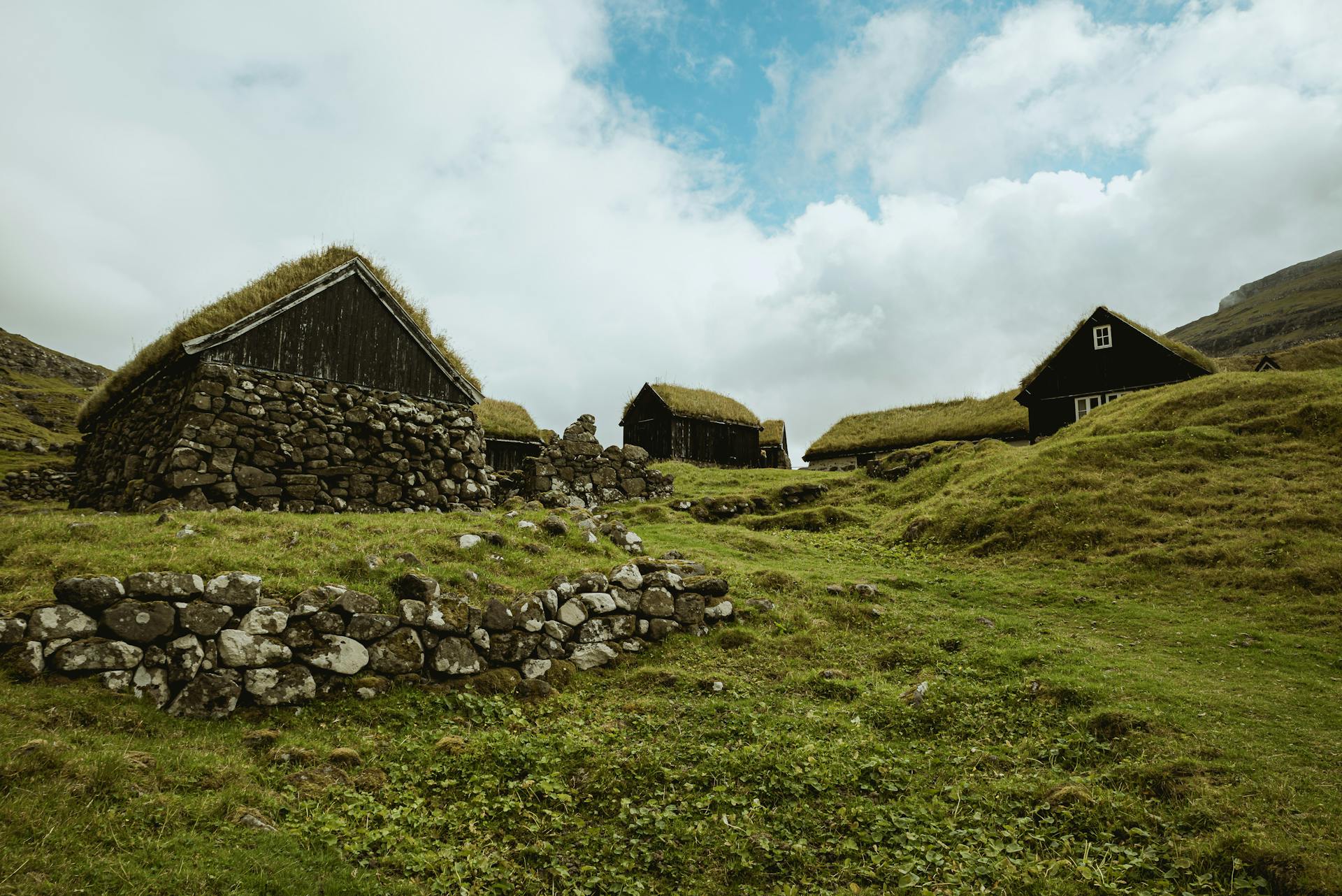 Stone houses with grass roofs in Sandoy, surrounded by lush greenery and a serene landscape under a cloudy sky.