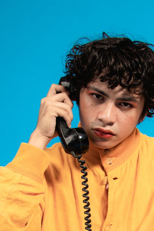 Close-Up Shot of a Man in Yellow Jacket Holding a Black Analog Phone on Blue Background