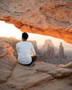 在美国犹他州日出时分，一名男子坐在峡谷地国家公园的梅萨拱门上