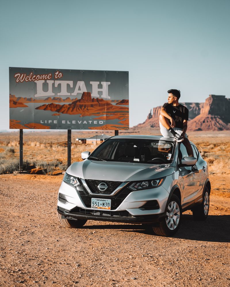 Young man sitting on a car by the Welcome to Utah sign in Monument Valley.