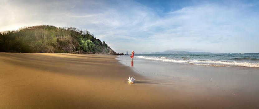 Distant female wearing red maxi dress and strolling on wet sandy seacoast against lush hilly cliff on sunny summer day