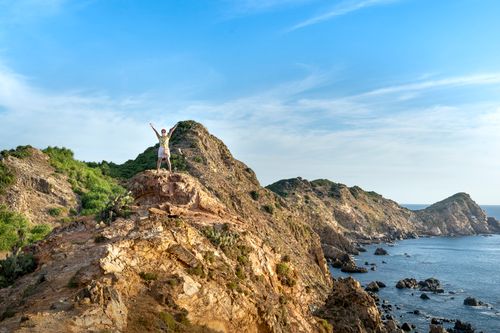 Free Full body unrecognizable person in summer outfit and sunglasses raising arms on rocky seashore washed by blue seawater under clear blue sky Stock Photo