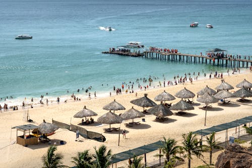 People relaxing on spacious sandy beach with straw umbrellas and wooden pier on sunny hot summer day