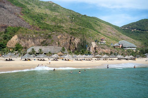 Scenic view of sandy beach with straw umbrellas washed by azure seawater and surrounded by rough grassy cliffs on hot summer day